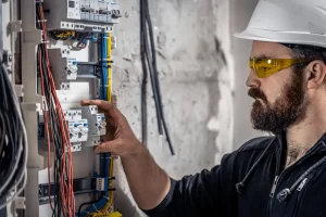 Electrician wearing safety glasses inspecting wiring and breakers inside a switchboard panel